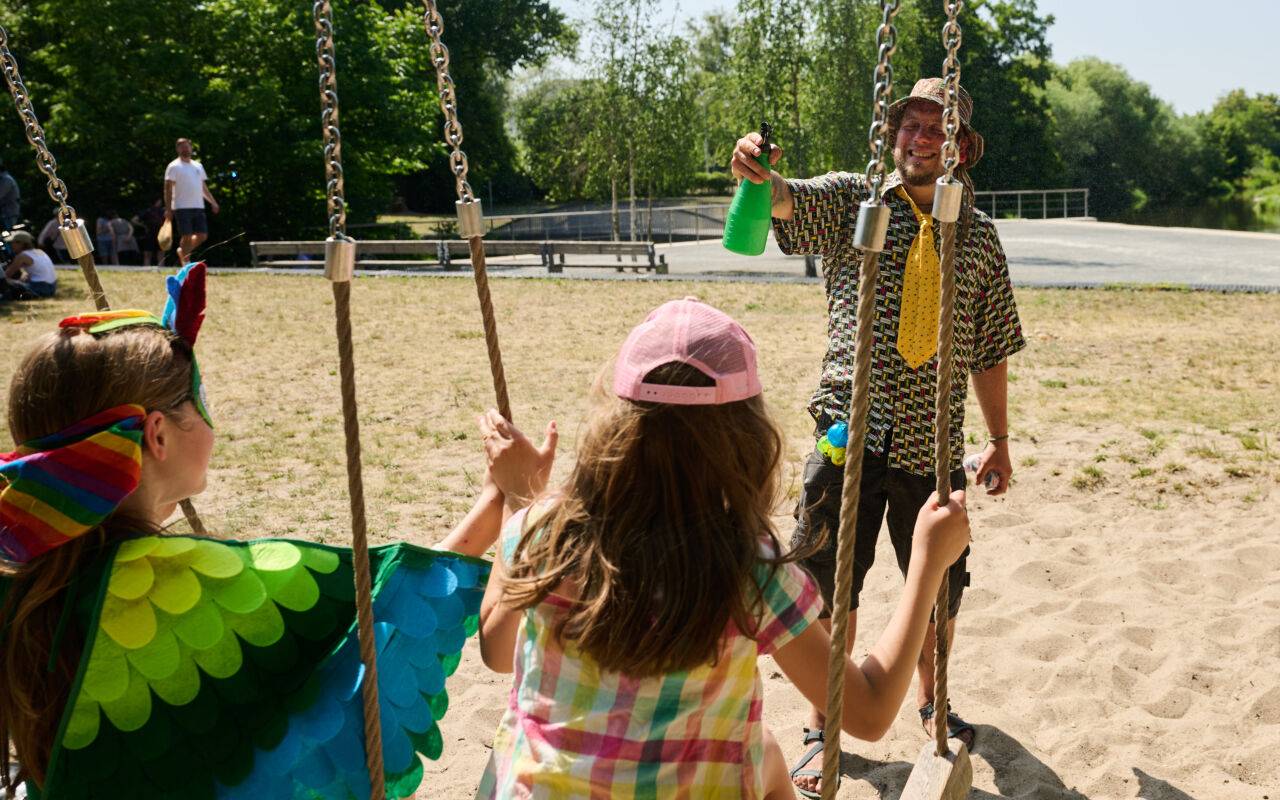 Ein Mann bespritzt seine Kinder mit Wasser, während diese auf Schaukeln sitzen. Zum Sommeranfang herrschen hochsommerliche Tempertauren.