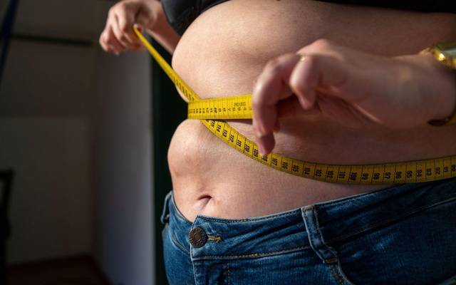 Eine Frau mit Übergewicht hält ein Maßband um ihren Bauch.